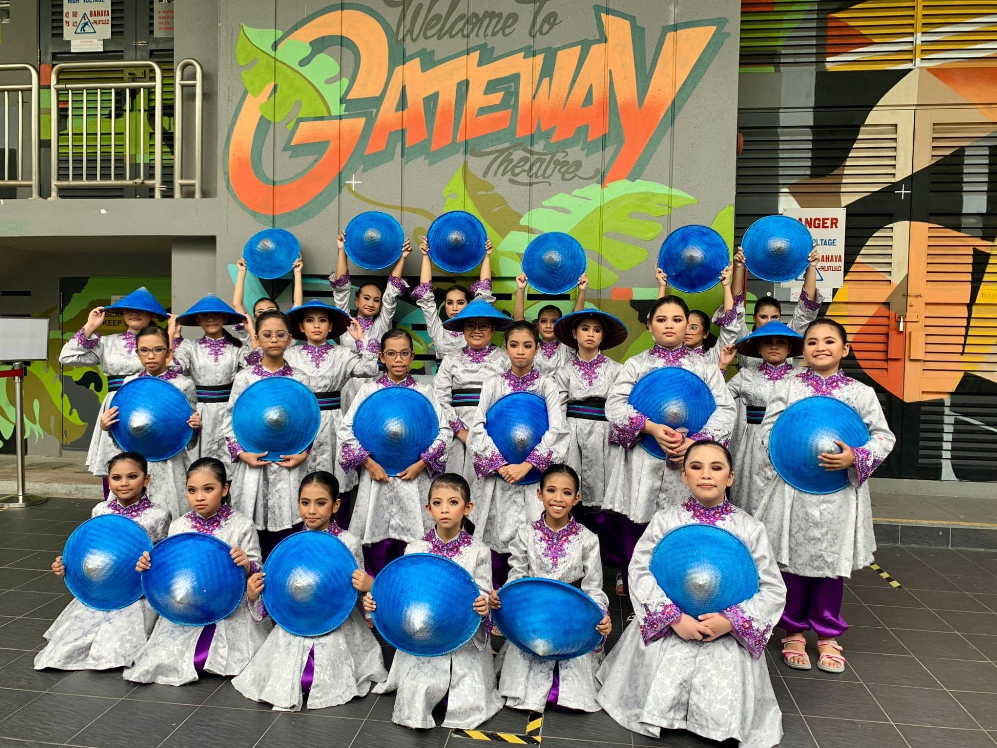 Group of children in silver outfits hold blue round hats in front of a mural reading Welcome to Gateway Theatre
